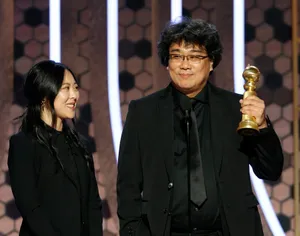 Sharon Choi and Bong Joon Ho standing onstage. They both wear all-black suits. Bong Joon Ho holds a Golden Globe trophy in his left hand.