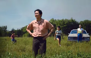 Steven Yeun stands in a field with his hands on his hips, wearing a short-sleeve salmon button-down shirt and brown pants. A woman stands behind him while two children run through the field.