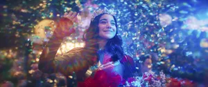 Iman Vellani as Kamala Khan smiles and waves, wearing her Ms. Marvel costume and a tiara and holding a bouquet of flowers as colored lights flash and confetti falls around her.