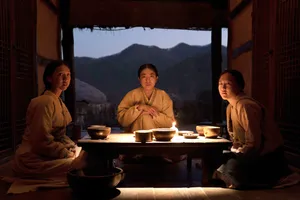 Three women sit around a candlelit table in a traditional Korean home, with an open wall behind them showing a view of the mountains.