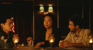 Nora sits at a dimly lit bar between Hae Sung and Arthur. The two men stare at each other while Nora looks straight ahead.