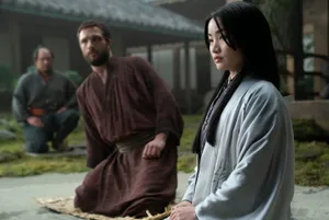 A bearded white man in a brown robe kneels on a woven mat in an outdoor courtyard. A Japanese woman sits next to him, wearing a gray kimono.