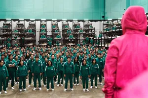 A crowd of people in numbered green tracksuits stand in a large room lined with tall bunk beds while a person in a hot pink sweatshirt addresses them.