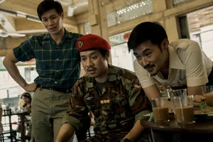 Hoa Xuande, wearing a blue plaid shirt, stands behind two men sitting at a table in an open-air restaurant. The man on the left wears military fatigues and the man on the right wears a white polo shirt. There are half-drunk glasses of coffee on the table.