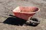 A red plastic wheelbarrow, covered in mud and muck, sitting in a dirt yard.