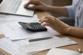 A stock photo of a person working on their taxes using a calculator and their laptop.