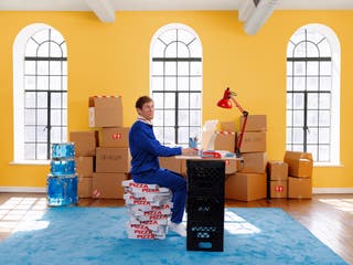 Piles of cardboard boxes bearing labels like "Bedroom" and "Dishes" sit in an empty, sunny room. In the foreground, a man in a blue tracksuit sits on a stack of pizza boxes, working at a makeshift desk made from milk crates.